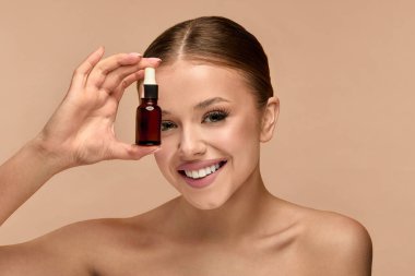 Kadın stüdyo ortamında gülümserken elinde serum gösteriyor. Cildinin sağlıklı cilde odaklanmasını ve basit kozmetik ürünler kullanarak ışık saçmasını teşvik ediyor..