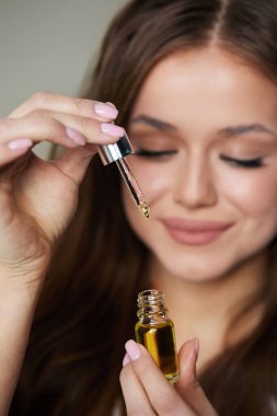 Genç bir kadın damlatıcıyı küçük bir şişenin üzerinde tutuyor ve yavaşça sıkıp gerekli yağı sürüyor. Odaklanmış ve huzurlu görünüyor, bir anlık özbakımı yansıtıyor..