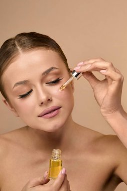 Bir kadın yüz yağını damlatıcıyla sürmeye hazırlanıyor, cildine iyi aydınlatılmış bir ortamda bakıyor. Gün ışığında günlük cilt bakımına odaklanmış ve meşgul görünüyor..