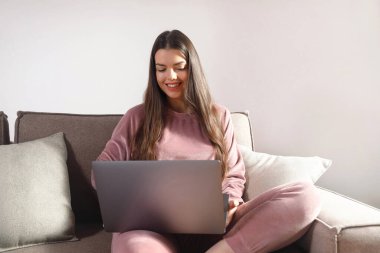 Gri bir koltukta oturan bir kadın dizüstü bilgisayarına odaklanmış. O, günlük giysiler giyer ve evdeki görevlerini tamamlarken neşeli bir ifade gösterir..