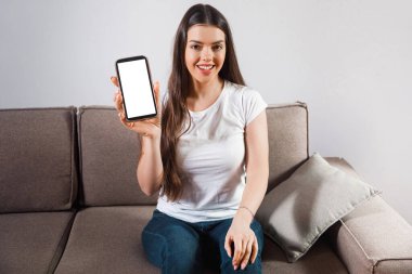 Parlak bir odada yumuşak bir koltukta oturan bir kadın. Elinde bir akıllı telefon tutuyor ve kameraya gülümsüyor. Telefon ekranı boş. Doğal ışık boşluğu doldurur..