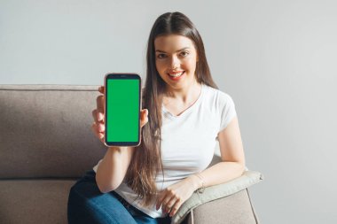 A woman sits on a sofa and holds up a smartphone with a blank screen. She smiles and looks at the camera. The setting is a living room with light coming in.