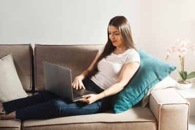 Evdeki bir kanepede oturan bir kadın dizüstü bilgisayarla çalışıyor. Arkasında bir yastıkla rahatlıyor. Etrafı ışıkla çevrili ve bir kaç kapalı masa bitkisiyle dolu..