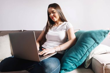 Bir kadın kanepede bağdaş kurup dizüstü bilgisayarında odaklanmış bir ifadeyle oturuyor. Parlak yastıklar gün boyunca rahat oturma odasına renk katar.