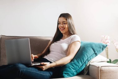 Bir kadın kanepede, dizüstü bilgisayarıyla gülümserken çalışıyor. Rastgele giyinmiş ve etrafında yumuşak bir yastık ve çiçekler var. Oturma odasında eğleniyor..