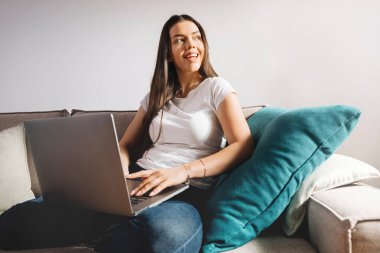 Önünde dizüstü bilgisayarı olan bir kadın kanepede oturuyor. Ekrana bakarken gülümsüyor. Yumuşak ışık pencereden girer ve odada parlak bir atmosfer yaratır..