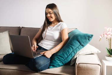 Bir kadın kucağında dizüstü bilgisayarla kanepede oturuyor. Çalışırken ya da evde dolaşırken mutlu görünüyor. Odanın açık renkleri ve konforlu yastıkları var..