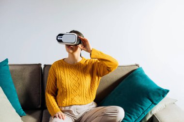 Bir kadın görsel içerikle meşgul olmak için ellerini hareket ettirirken sanal gerçeklik kulaklığı kullanıyor. Işıklı duvarları ve basit bir geçmişi olan bir odada..