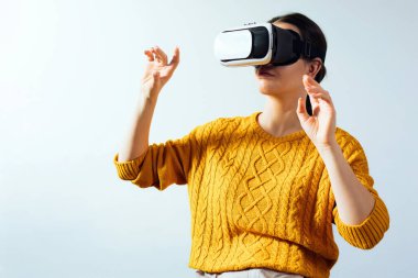 A woman is using a virtual reality headset while moving her hands to engage with the visual content. She is inside a room with light walls and a simple background.