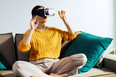 Bir kadın görsel içerikle meşgul olmak için ellerini hareket ettirirken sanal gerçeklik kulaklığı kullanıyor. Işıklı duvarları ve basit bir geçmişi olan bir odada..