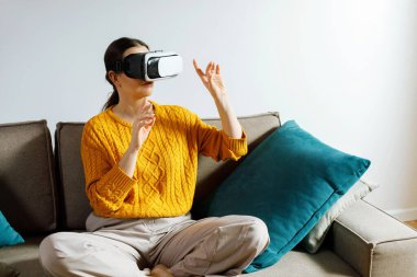 Evdeki koltukta bağdaş kurmuş bir kadın sanal gerçeklik kulaklığı takıyor. Dijital bir deneyimle etkileşime girerek ellerini kaldırıyor..