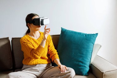 Sanal gerçeklik kulaklığı takmış bir kadın kanepeye sessizce oturur. Gün boyunca oturma odasındaki sanal deneyimle meşgul ve odaklanmış görünüyor..