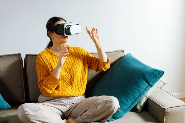 Evdeki koltukta bağdaş kurmuş bir kadın sanal gerçeklik kulaklığı takıyor. Dijital bir deneyimle etkileşime girerek ellerini kaldırıyor..