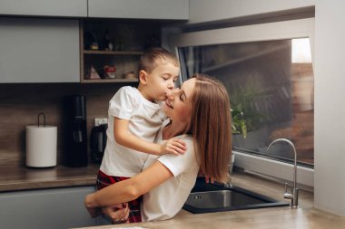 Anne, oğlunu bir mutfakta tutuyor ve keyifli bir anı paylaşıyorlar. Güneş ışığı pencereden girer, birlikte kahvaltı yaparken yüzlerini aydınlatır..