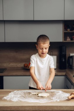 A young child works with dough on a kitchen counter, using hands to shape it. The setting includes light-colored cabinets and a few kitchen tools nearby. Fresh ingredients are visible.