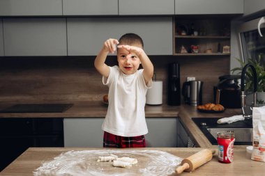 Çocuk mutfakta durur ve tezgahta hamur sarar, yemek hazırlarken heyecanını gösterir. Sabah saatlerinde çeşitli mutfak aletleri ve malzemeler mevcut..