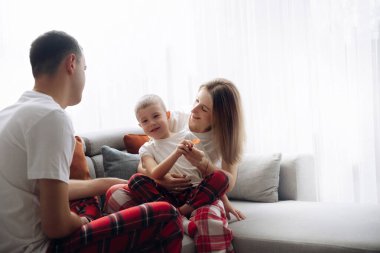 Bir aile sabahları kanepede birlikte rahat vakit geçirir. Çocuk aynı pijamaları giyen ebeveynler arasında otururken gülümsüyor..