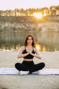 Güneş batarken gölün kenarındaki minderde yoga yapan bir kadın huzurlu bir atmosfer yaratıyor. Duruşu odak ve uyumu gösteriyor..