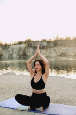 Siyah bluzlu bir kadın gün batımında gölün kenarında yoga yapıyor. Kayalıklar suya yansıyarak, barışçıl bir atmosfer yaratıyor..