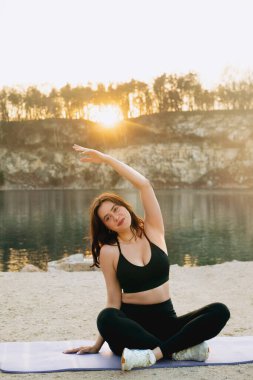 A young woman is engaged in yoga, sitting on a mat by the water. She stretches her arm upward as the sun sets behind the trees, creating a serene scene.
