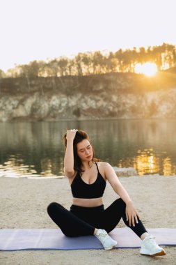 Gün doğumunda dingin bir gölün kenarındaki minderde yoga yapan bir kadın. Dengesini huzurlu bir ortamda bulduğunda altın ışık suda parıldıyor..