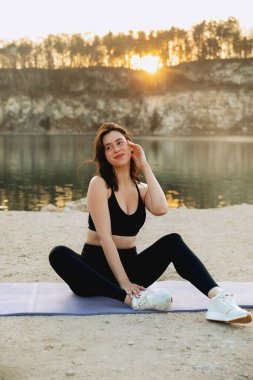 Genç bir kadın gün doğumunda göl kenarında yoga yapıyor. Etrafı sakin su ve ağaçlarla çevrili. Doğanın güzelliğinde rahatlık ve farkındalığı teşvik ediyor..