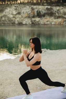 Bir kadın, güneşli bir günde, güzel uçurumlar ve yeşilliklerle çevrili sakin bir gölde yoga minderinde hamlesini yapıyor..