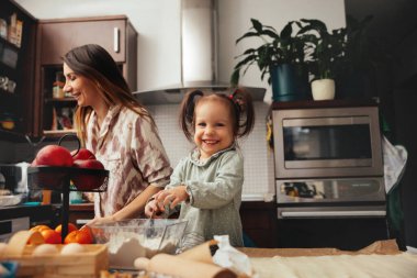 Bir kız, annesinin mutfakta pişirmesine yardım ederken gülümser. Etrafları meyvelerle ve pişirme aletleriyle çevrili, birlikte vakit geçirmekten zevk alıyorlar..