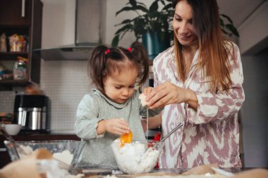 Bir kadın, küçük kızının yumurta kırarak karıştırmasına yardım eder. Mutfakta bir anlarını paylaşıyorlar, etrafları malzemelerle ve yemek aletleriyle çevrili, birlikte vakit geçiriyorlar..