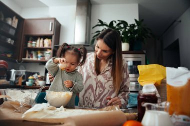 Bir anne ve küçük kızı, parlak bir mutfakta kurabiye pişirirken malzemeleri bir kasede karıştırıyorlar. Birçok malzeme ve alet tezgahta, eğlenceli aktivitelerini gösteriyor..