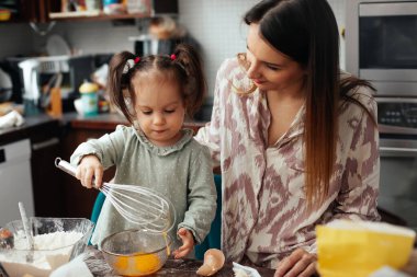 Bir anne ve kızı mutfakta birlikte çalışıyorlar, hamuru çırpıcıyla karıştırıyorlar. Güneşli bir öğleden sonra pişirmeye hazırlanırken eğleniyorlar..