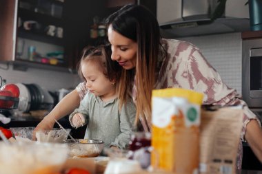 Bir anne ve kızı mutfakta tatlı hazırlamakla meşguller. Tezgahta çeşitli pişirme malzemeleriyle çevriliyken malzemeleri bir kasede karıştırıyorlar..