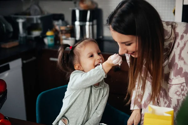 Bir çocuk mutfakta annesine gülümserken eline un tutarak uzanır. Mutfak eşyalarıyla çevrili eğlenceli bir yemek pişirme anından hoşlanıyor gibiler..