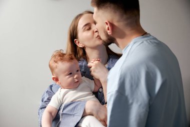 İki ebeveyn bebeklerini tutarken nazikçe öpüşürler. Sahne yumuşak renklerle sıcak bir ev atmosferi gösteriyor. Bebek her iki ebeveyne de merakla bakıyor..