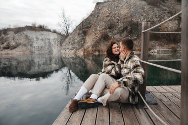 İki insan, bulutlu bir günde gülümseme ve şefkat paylaşırken sakin bir su kütlesinin yanında rahatlar.