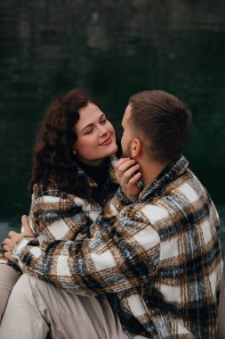 Bir erkek ve kadın, suyun kenarındaki sakin bir noktada birlikte oturur. Özel bir günde sıcak kıyafetler giyerek gülümseyerek ve nazik dokunuşlar yaparlar..