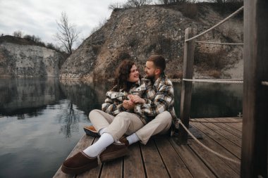 İki kişi sakin suyun yakınındaki ahşap bir rıhtımda oturuyor. Sevgililer Günü 'nde el ele tutuşup birbirlerine bakıp gülümsüyorlar..