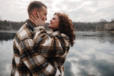 İki insan göl kenarında durup gülümseyerek birbirlerine bakıyorlar. Aynı süveteri giyip birlikte vakit geçirmekten hoşlanıyorlar..