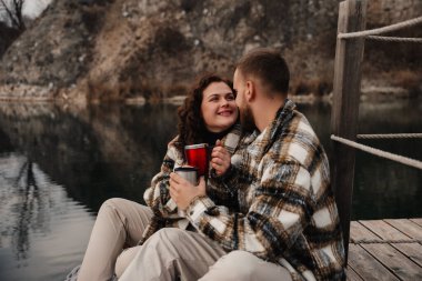 İki insan, gölün kenarında oturmuş, romantik bir ortamda gülümseyip bir anı paylaşırken ılık içeceklerden hoşlanırlar..