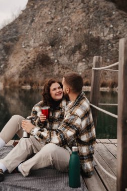 İki kişi rıhtımda yan yana oturur. Kadın ve erkek bardak tutarken gülümsüyor, su kenarında birbirlerine eşlik ederek eğleniyorlar..