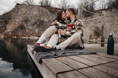 İki insan sakin bir gölün kıyısındaki iskelede rahatlar, içkilerini paylaşır ve sevgililer gününde romantik bir anın tadını çıkarırlar..