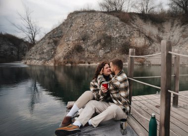 İki kişi rıhtıma yakın oturuyor, gülümsüyor ve bardaklarını tutuyor. Sevgililer Günü 'nde manzaralı bir ortamda bir anı paylaşıyorlar..