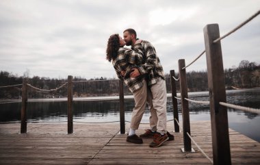 Tahta bir iskelede iki kişi öpüşüyor. Bulutlu bir gökyüzünün altında ağaçlarla çevrili durgun suyun yanındalar..