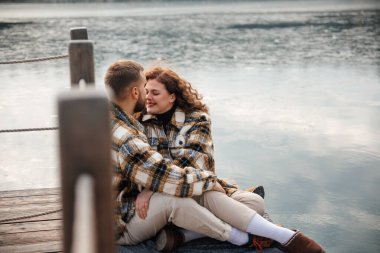 İki insan, sevgililer gününde sıcacık giysilerle sarılmış bir şekilde rıhtıma yakın oturur, gülümser ve sevgisini gösterir..