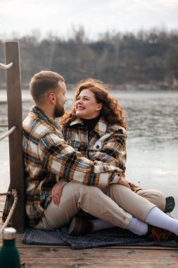 Sevgililer Günü 'nde eşleşen ceketler giyerek rıhtımda oturmuş birbirlerine gülümseyen bir erkek ve kadın birlikte vakit geçiriyorlar.