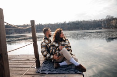 İki insan göl kıyısındaki bir rıhtımda kalın ceketler giyerek ve bir battaniyeye oturup eğlenirken birbirlerine eşlik ederler..