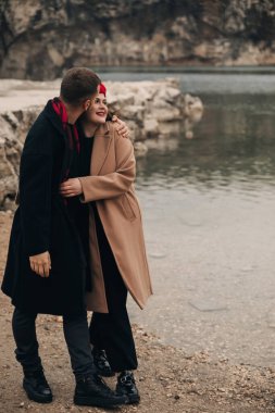 İki insan denize yakın durur, sevgililer gününde birbirlerine sarılır ve gülümserler. Manzaralı bir yerde..