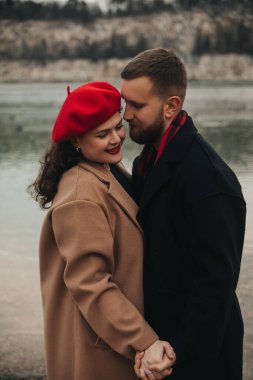 Bir erkek ve kadın, deniz kenarında sevgi dolu bir kucaklaşmayı paylaşıyor. Kış için sıcak kıyafetler giyerken el ele tutuşup birbirlerine gülümserler..