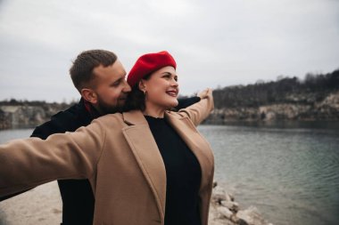 Kadın kollarını açarken bir erkek ve bir kadın suyun yanında durup gülümsüyor. Serin bir günde neşe ve bağlantı anlarını paylaşıyorlar..