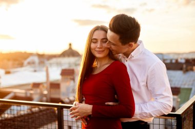 Bir erkek ve bir kadın, güneş batarken bir çatıda kucaklaşmanın keyfini çıkarırlar. Şehir manzarasına sıcak bir ışık tutarlar..